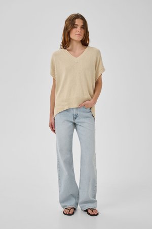 Femeie purtând un pulover bej fără mâneci cu decolteu în V, blugi largi albastru deschis și șlapi negri, stând cu o mână în buzunar.