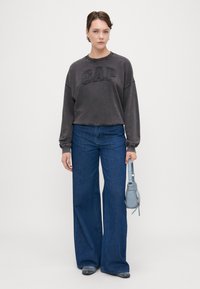 Sudadera gris recortada con el logo "GAP", combinada con unos jeans de pierna ancha azules. Textura ligera en la tela, sosteniendo un pequeño bolso azul.