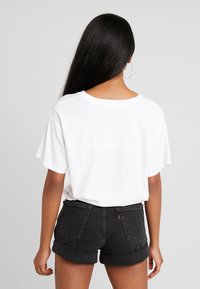 Camiseta blanca de gran tamaño con mangas cortas, combinada con pantalones cortos de mezclilla negra. Detalle de costura visible en la parte trasera y dobladillos enrollados en los pantalones cortos.
