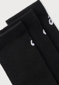 Μαύρες ριγωτές κάλτσες πλήρους μήκους διπλωμένες δίπλα-δίπλα με μερική λευκή λεπτομέρεια λογότυπου κοντά στον μανσέτα σε λευκό φόντο.