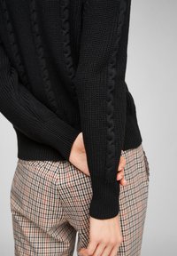 Une personne portant un pull en maille torsadée noir et un pantalon à carreaux, tenant d'une main le poignet opposé derrière le dos.