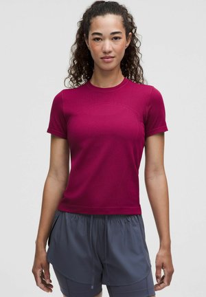 Jeune femme aux cheveux bouclés portant un haut magenta manches courtes et un short ample gris foncé, debout devant un fond uni.