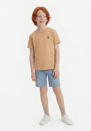 Rothaariger Junge lächelt, trägt ein hellbraunes kurzärmeliges Hemd mit Schmetterlingslogo, hellblaue Jeans-Shorts, beigetöne Socken und weiße Turnschuhe, steht.