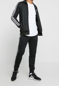 Musta urheiluasu, jossa on valkoiset raidat hihoissa ja housuissa, sisältäen vetoketjullisen takin ja kapea jalkatyylisiä housuja.