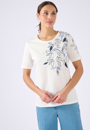 Femme portant un haut blanc à manches courtes avec des motifs de feuilles bleues, roses et vertes, associé à un pantalon bleu clair, sur un fond blanc.