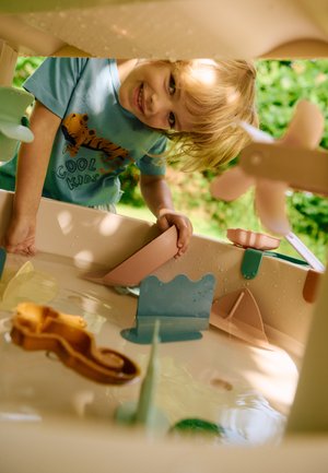 Plastikk vannleke bord i lys beige med fargerike tilbehør: rosa, blå og gule former; teksturerte overflater; vanndråper på kantene.