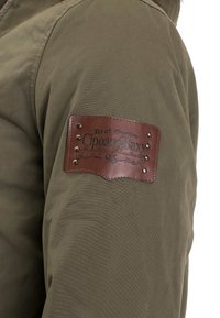 Olivenfarbene Stoffjacke mit einem braunen Lederpatch, auf dem der Text "Real Denim CipoandBaxx" steht, sowie dekorativen Metallnieten.