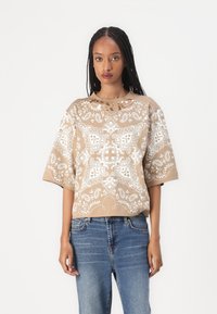 Beige t-skjorte med kort erme og hvitt paisley-mønster, med rund hals og avslappet passform. Brukt sammen med blå denimbukser.
