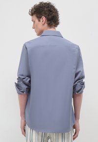 Mann mit lockigem Haar, der ein hellblaues Langarmhemd mit hochgekrempelten Ärmeln und gestreiften Hosen trägt, mit dem Rücken zu einer neutralen Hintergrund.