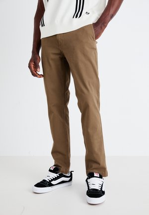 Khaki trousers made of cotton with a straight fit, worn with black and white sneakers. Notable stitching details on the shoes.