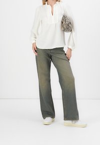 Biała bluzka z długim rękawem i falbanistym dekoltem, zestawiona z szerokimi, jasnoniebieskimi dżinsami i białymi trampkami. Trzyma torebkę w wężowy wzór o teksturowanej powierzchni.