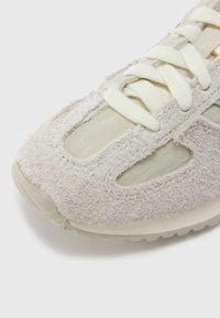 Pantof sport gri deschis cu partea superioară din piele întoarsă texturată, secțiuni din plasă și șireturi crem. Are o talpă din cauciuc cu decupaje și vârf rotund.