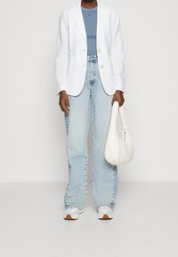 Blazer blanco de lino sobre una camiseta de cuello redondo azul, combinado con jeans acampanados de un azul claro y un bolso texturizado blanco. Unas zapatillas blancas completan el look.
