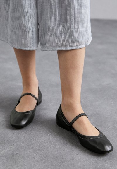 Blöße Beine in schwarzen Leder-Mary-Jane-Ballen mit gewellten Knöchelriemen und hellgrauen, cropped Hosen auf einem grauen Boden.