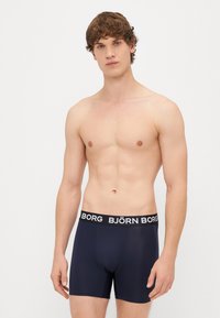 Młody mężczyzna z jasną skórą i kręconymi włosami, ubrany w granatowe majtki bokserki z czarnym pasem marki "BJÖRN BORG", stojący na tle białym.