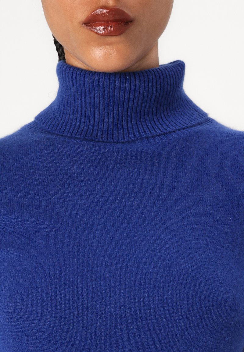 Femme portant un pull col roulé côtelé bleu royal avec un rouge à lèvres marron brillant, visible de la mâchoire aux épaules sur fond blanc.