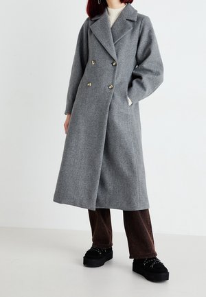 Kobieta ubrana w długi, szary dwurzędowy płaszcz, ciemnobrązowe spodnie, czarne buty na platformie i biały sweter, stojąca na tle białego tła.