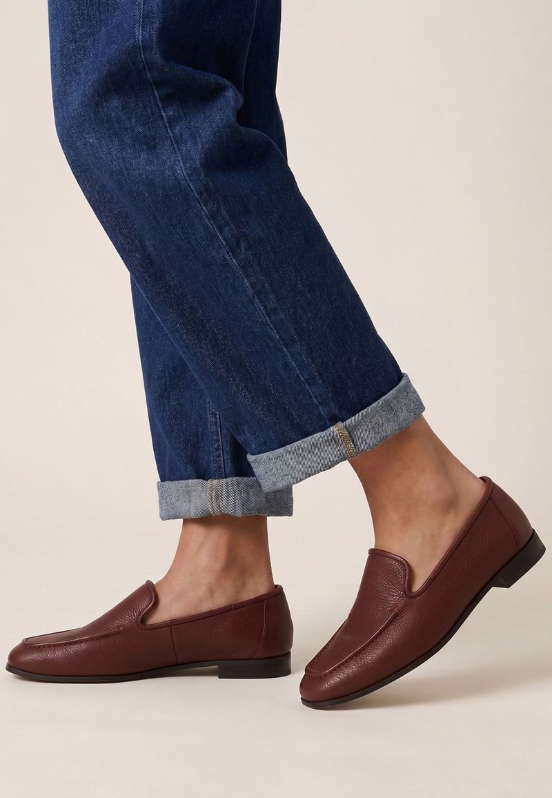 Person iført brune læderslip-on sko med mørkeblå opkørte denimjeans på en lys baggrund.