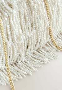 Bílá kabelka s třásněmi z korálků a zlatým řetízkovým popruhem. Korálky jsou kulaté a lesklé, vytvářející texturovaný vzhled.