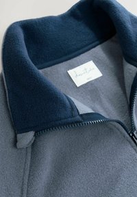Fleece jack met een marineblauwe kraag en een grijs lijf. Heeft een rits en een 'avontuur' label aan de binnenkant. Zachte textuur en gelaagd ontwerp.