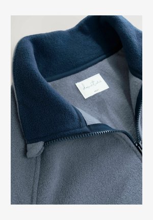 Fleece bunda s námorníckym golierom a šedým telom. Obsahuje zips a vnútornú nálepku s nápisom 'adventure'. Mäkká textúra a vrstvený dizajn.