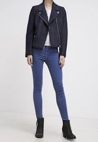 Kobieta w czarnej, teksturowanej kurtce na zamek, białej koronkowej bluzce, niebieskich skinny jeansach i czarnych botkach do kostki, stojąca na tle gładkiego białego tła.