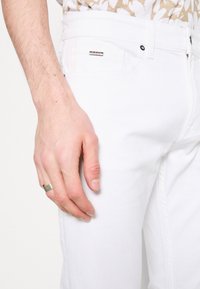 Gros plan d'une main avec une bague en argent reposant à côté d'une poche de pantalon blanc avec une petite étiquette de marque visible.