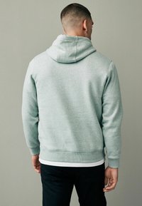 Lichtgrijze hoodie van een gestructureerde stof, met verstelbare capuchon en geribbelde boorden. Draagt over een wit shirt, gecombineerd met een zwarte broek.
