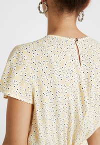 Robe jaune clair avec un motif floral de points blancs et noirs, manches courtes et fermeture en clé dans le dos avec un détail de bouton.