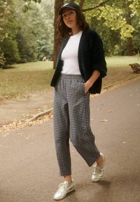 Groene textuur cardigan over een witte t-shirt, blauw en witte geruite broek en zilveren sneakers, wandelend op een pad in een park.
