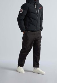 Veste matelassée noire avec fermeture éclair à l'avant, ornée d'un écusson logo sur la manche. Associée à un pantalon cargo foncé et des baskets blanches.