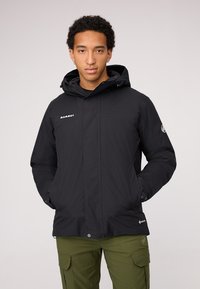 Giacca impermeabile nera con cappuccio, colletto alto e zip frontale; presenta un logo sul petto e sul lato sinistro in basso. Indossata con pantaloni cargo verdi.
