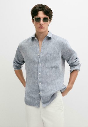 Ung mand iført runde solbriller, en lyseblå linned langærmet skjorte med opkrogede ærmer og hvide bukser, stående med hænderne i siden.