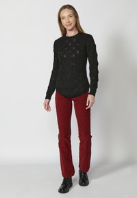 Czarny sweter dzianinowy z ażurowym wzorem, zestawiony z czerwonymi spodniami kordurowymi. Modelka nosi czarne botki na kostkę; tło jest jednolicie białe.