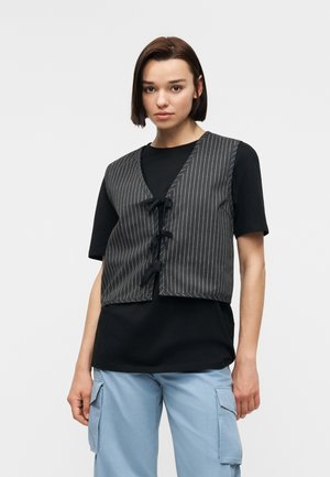 Frau mit glattem braunem Haar trägt ein schwarzes Kurzarmshirt, eine graue Nadelstreifenweste mit Bändern und hellblaue Cargohosen, steht vor weißem Hintergrund.