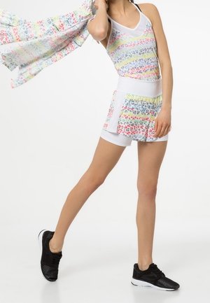 Vrouw draagt een meerkleurige gestreepte sportoutfit met short en tanktop, houdt een bijpassende patroonstof in beweging, zwarte sneakers op een witte vloer.