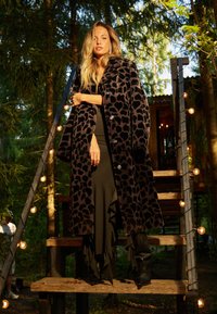 Capot lung negru cu imprimeu de leopard, textură moale, butoane mari, purtat peste o rochie întunecată cu volane, asortat cu cizme negre mulate.