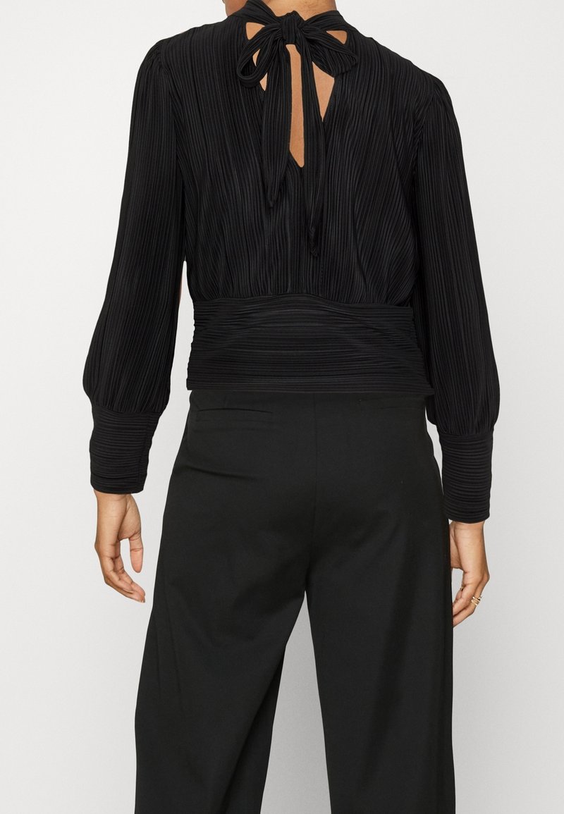 Femme portant un chemisier plissé noir à manches longues avec un nœud au dos, associé à un pantalon noir taille haute.