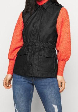 Gilet nero senza maniche con colletto, vita elastica, bottoni a pressione frontali e due tasche a toppa, indossato sopra un maglione rosso brillante.