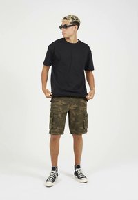 Juoda medvilninė marškinėlių su laisvu siluetu; kamufliažinės cargo šortai su daugybe kišenių; juodi sportbačiai; modelis dėvi tamsius akinius nuo saulės.