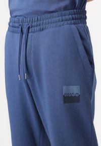 Mornarsko modri trenirki z elastiko v pasu, vrvico in stranskimi žepi. Ponuja HUGO logotip v temnejši odtenek. Mehka tekstura.