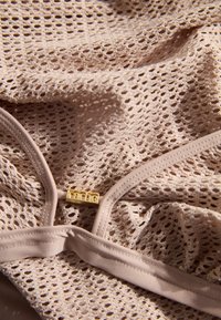 Tissu en dentelle rose clair avec un motif en maille, comportant une étiquette logo dorée et des bords lisses. La forme et la texture sont douces et délicates.