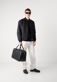Schwarze Bomberjacke über einem schwarzen Shirt, weiße Hose, schwarze Tasche mit zwei Griffen und schwarze Schuhe. Das Model trägt eine dunkle Sonnenbrille.