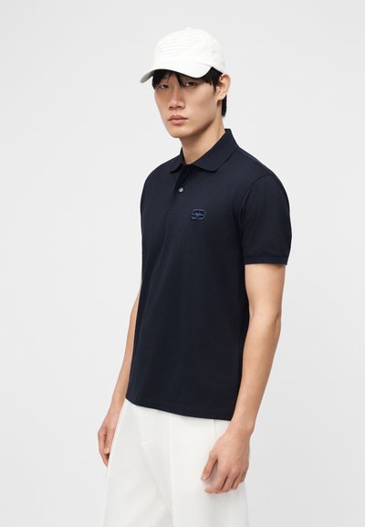 Άνδρας που φοράει μαύρο πόλο μπλουζάκι με κοντά μανίκια και μικρό λογότυπο, λευκό παντελόνι και λευκό καπέλο μπέιζμπολ, στέκεται μπροστά σε απλό φόντο.