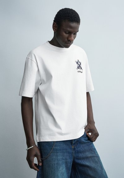 Jeune homme portant un t-shirt blanc Off-White avec un jean et un accessoire en chaîne, regardant vers le bas sur un fond uni gris clair.