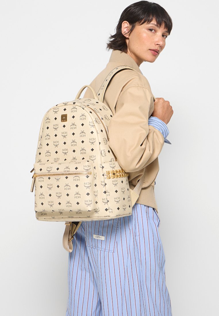 Beiger Leder-Rucksack mit einem sich wiederholenden Logo-Muster, goldfarbenen Beschlägen und einer Front-Reißverschlusstasche. Verstellbare Träger und abgerundete Form.