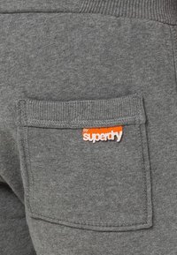 Detalle de pantalones deportivos de algodón gris con un bolsillo parche con la marca "superdry" en naranja y blanco, y puño acanalado texturizado en la abertura.