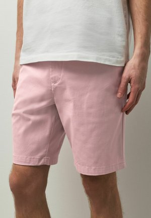 Mann trägt hellrosa knielange Shorts und ein weißes Hemd, steht mit entspannten Händen an den Seiten vor einem einfarbigen Hintergrund.