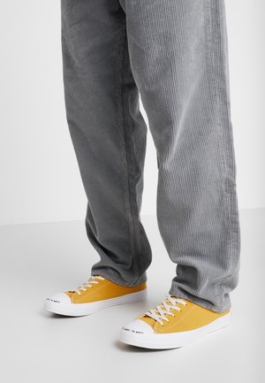 Zapatillas de lona amarilla con suelas y punteras de goma blanca, diseño con cordones y detalle de texto. Combinadas con pantalones de pana gris.