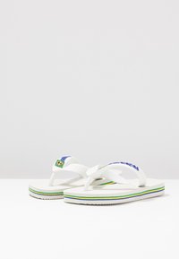 Par hvide flip-flops med blå, grønne og gule striber på sålerne og et lille brasiliansk flag på stropperne.